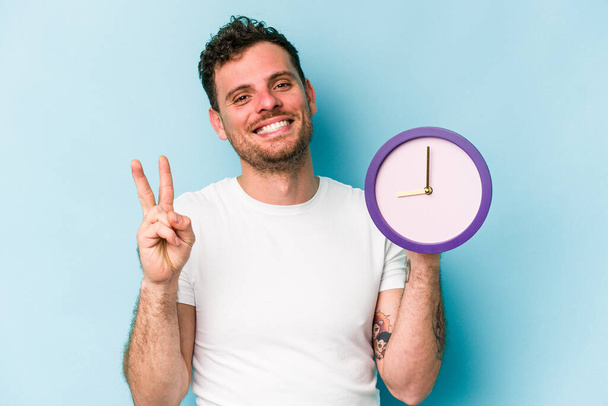 Young caucasian man holding a clock isolated on blue background joyful and carefree showing a peace symbol with fingers. - Fotografie, Obrázek