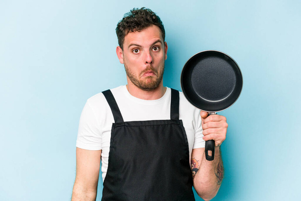 Молода кавказька плита тримає сковороду ізольовану на синьому фоні раковини плечі і відкриті очі збентежені
. - Фото, зображення