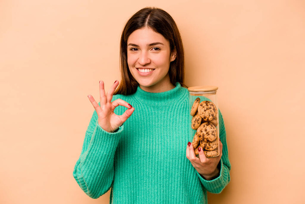 Young hispanic woman holding cookies jar isolated on beige background cheerful and confident showing ok gesture. - Photo, Image