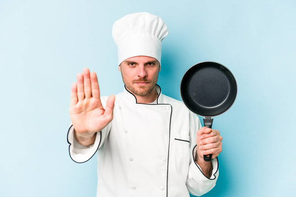 Young caucasian chef man holding flying pan isolated on blue background standing with outstretched hand showing stop sign, preventing you. - Photo, Image