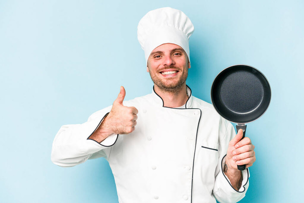 Young caucasian chef man holding flying pan isolated on blue background smiling and raising thumb up - Foto, imagen
