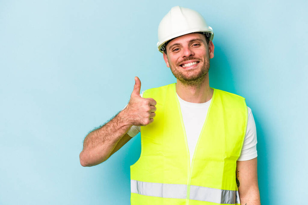 Genç işçi beyaz adam mavi arka planda izole edilmiş gülümsüyor ve başparmağını kaldırıyor. - Fotoğraf, Görsel