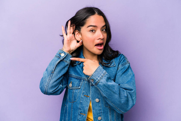 Young hispanic woman isolated on purple background trying to listening a gossip. - 写真・画像