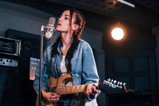 Young beautiful female performer with guitar singing and rehearsing in a recording studio. - Foto, imagen