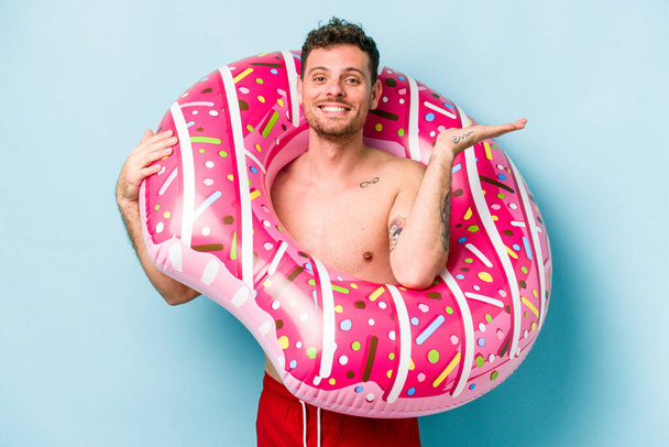 Young caucasian man holding an inflatable donut isolated on blue background showing a copy space on a palm and holding another hand on waist. - Photo, image