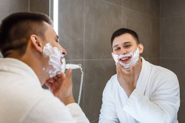 Чоловік у білій шубці з піною для гоління на обличчі. Відображення у дзеркалі, чудовий настрій
. - Фото, зображення