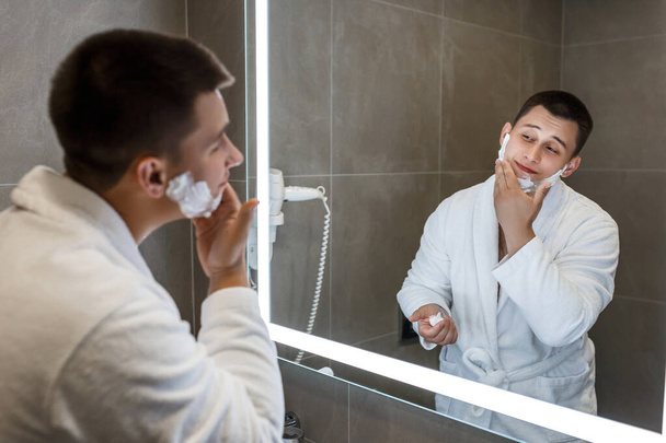 A man in a white coat with shaving foam on his face. Reflection in the mirror, great mood. - Photo, Image