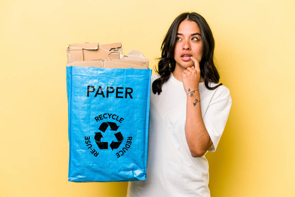 Jovem hispânica reciclando papel isolado no fundo amarelo relaxado pensando em algo olhando para um espaço de cópia. - Foto, Imagem