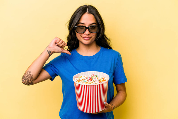 jonge Spaanse vrouw houden popcorn geïsoleerd op gele achtergrond voelt trots en zelfvertrouwen, voorbeeld te volgen. - Foto, afbeelding