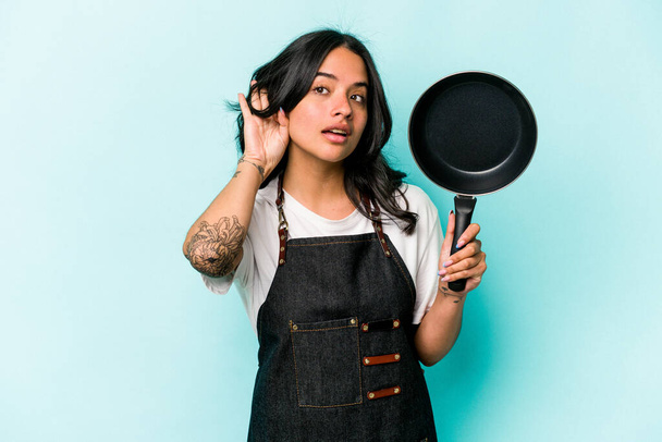 Joven cocinera hispana sosteniendo sartén aislada sobre fondo azul tratando de escuchar un chisme. - Foto, imagen
