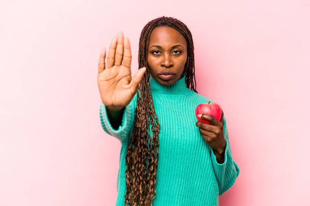 Fiatal afro-amerikai nő kezében egy alma elszigetelt rózsaszín háttér álló kinyújtott kézzel mutató stop jel, megakadályozza, hogy. - Fotó, kép