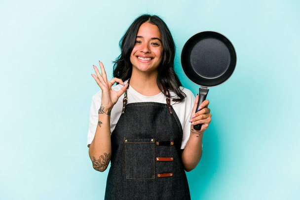 Joven cocina hispana mujer sosteniendo sartén aislado sobre fondo azul alegre y confiado mostrando gesto ok. - Foto, imagen
