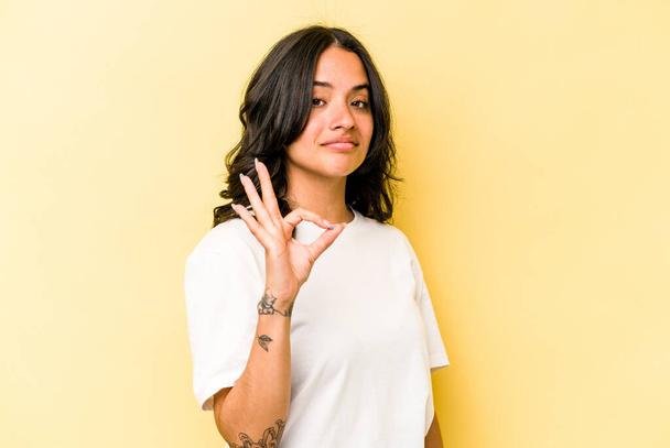 Junge hispanische Frau auf gelbem Hintergrund zwinkert mit einem Auge und hält eine Okay-Geste mit der Hand. - Foto, Bild