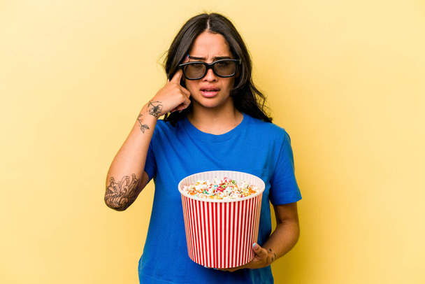 Young hispanic woman holding popcorn isolated on yellow background showing a disappointment gesture with forefinger. - Foto, afbeelding