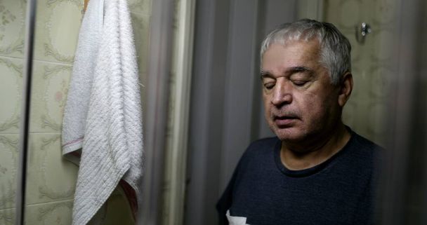 Old sad 60 year old man closing eyes in front bathroom mirror in reflection - Foto, afbeelding