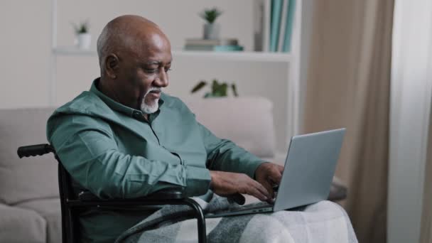 Old 60s african business man in wheelchair indoor at home typing on laptop communicates remotely working online distant texting chatting with family grandfather with disability use modern computer app - Footage, Video
