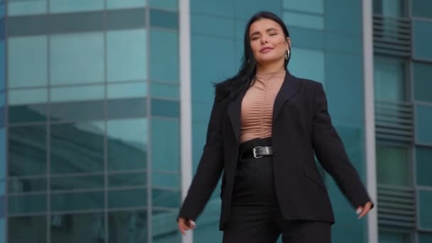 Young successful elegant hispanic businesswoman in business suit stands in confident pose with arms crossed against office building in background feeling success freedom achievement posing outdoors - Footage, Video