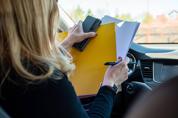 middle aged blonde business woman in glasses in car working with documents, rear view - Foto, Imagen