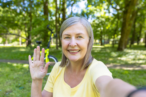 Senior gray-haired woman, active sportswoman in the park in the summer looks at the phone camera and smiles waving her hand at a video call - Foto, immagini