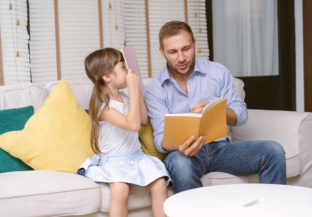 Jóképű apa ül a kanapén és mesekönyvet olvas a lányának otthon a nappaliban. Mosolygó egyedülálló apa gondoskodó kislány élvezi időt tölteni otthon. - Fotó, kép