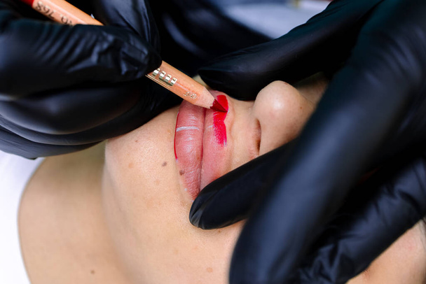 close-up of a permanent makeup master holding a red pencil and painting over the model's lips to perform a tattoo - Фото, зображення
