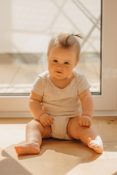 Uma menina gentil de 7 meses está sentada perto de uma porta da varanda em um bodysuit em casa - Foto, Imagem