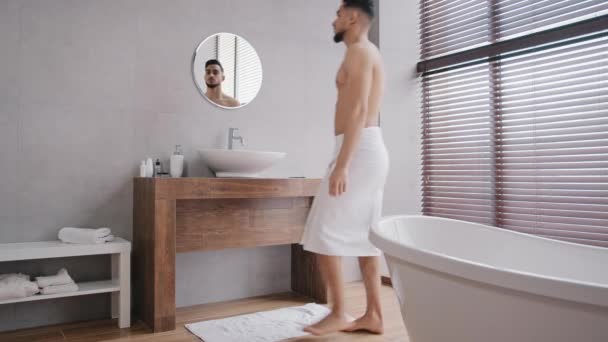 Naked bare sexy muscular Arab Indian man enters going walking in bath after shower with white towel on hips washes face with warm water in bathroom sink looks at reflection in mirror morning hygiene - Кадры, видео