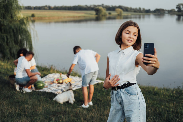 Adolescente caucásica chica haciendo selfie en el teléfono inteligente mientras su familia en el fondo teniendo picnic junto al lago al aire libre - Foto, Imagen