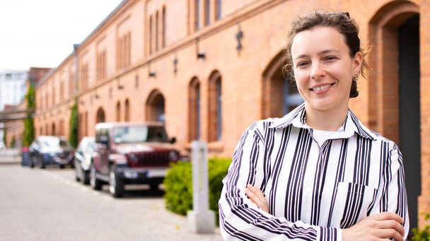 Young beautiful business woman in white black striped shirt smiling looking at camera, standing outdoors on red brick restaurant building, parked cars, green bush background. Build career with us. - Photo, image