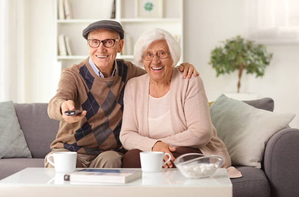 Happy elderly couple sitting on a sofa at home holding a remote control and watching tv - Photo, Image
