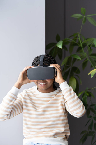 jong verbaasd enthousiast afro meisje dragen VR helm ontspannen met virtual reality op vensterbank thuis - Foto, afbeelding
