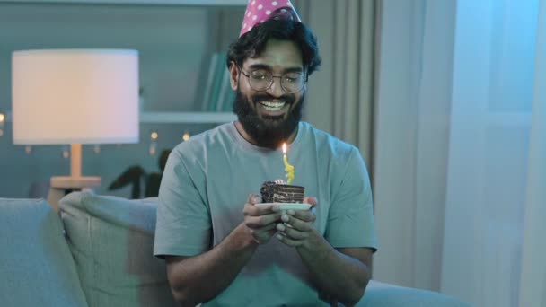 Дім веселий арабський індіанець бородатий чоловік в окулярах з капелюхом тримаючи на день народження шматочки торта зі свічкою, змушуючи бажати говорити про майбутню мрію, бажаючи підірвати день самотності сидячи на дивані - Кадри, відео