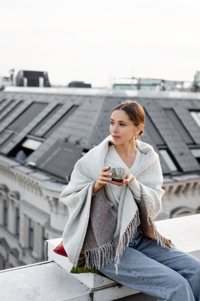 woman holding clay cup with tea while sitting on rooftop under shawl - Foto, afbeelding