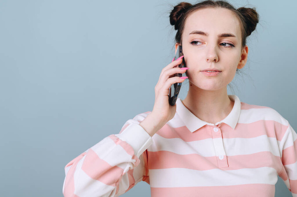 A girl with a funny hairstyle calls on the phone with an incredulous expression on her face. Crazy girl with a phone on a blue background - Фото, изображение