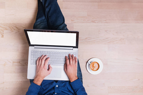 Man wearing jeans sitting on the wooden floor and holding laptop with a white empty screen - Photo, image
