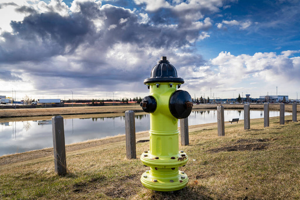 Airdrie Albertaの工業団地の嵐の保持池の隣に立つインフラストラクチャに接続された黄色の消火栓劇的な空の下でカナダ. - 写真・画像