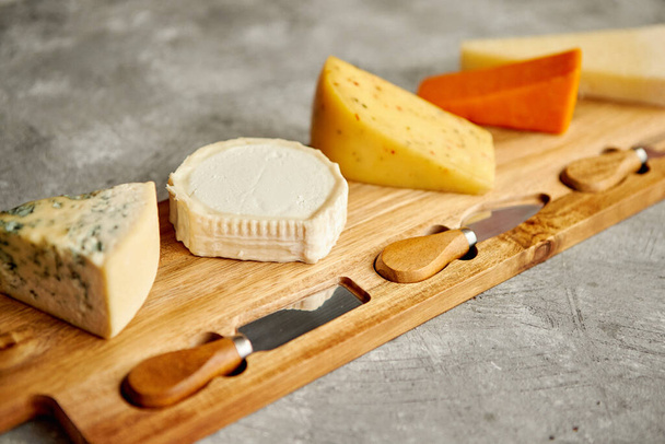 Assortment of various kinds of cheeses served on wooden board with fork and knives - Photo, Image