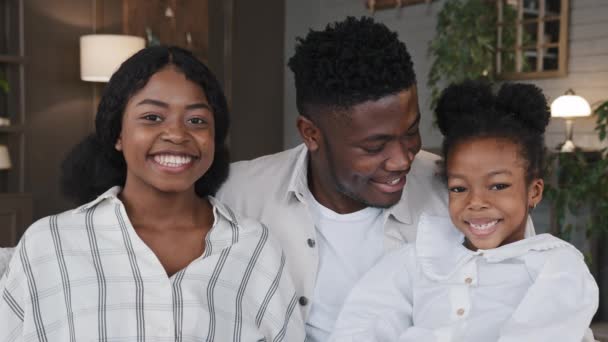 Gelukkig Afrikaans familieportret. Ouders met vrolijke dochter meisje kijken naar camera lachen knuffelen thuis glimlachende tanden. Positieve moeder, vader en kind knuffelen op de bank. Tandheelkundige gezondheidszorg - Video