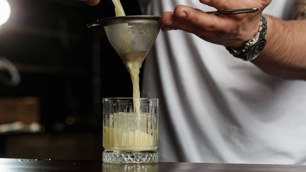 man bartender carefully filters bright alcoholic cocktail from steel shaker cup into glass through sieve. Close-up view - Foto, afbeelding