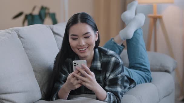 Asiatische weibliche entspannende Mädchen Frau auf der Couch zu Hause genießen Chat-Tippen App Handy entspannen Virales Live-Videospiel ansehen folgen wie Beitrag in sozialen Medien teilen und Lächeln Emoji scrollen Web senden - Filmmaterial, Video