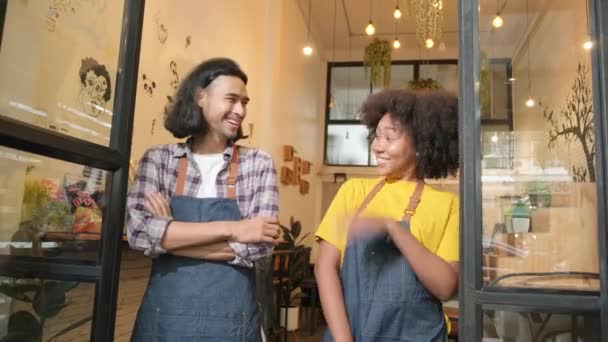 Two young startup barista partners with aprons stand at casual cafe door, arms crossed, laugh and tease together, happy and cheerful smiles with coffee shop service jobs, small business entrepreneurs. - Кадры, видео