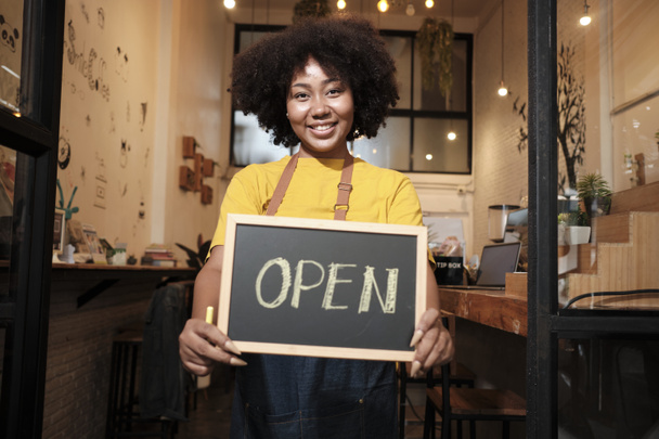 One African American female startup barista stands at casual cafe door, looks at camera, and shows open sign, happy and cheerful smiles with coffee shop service jobs, and new business entrepreneurs. - Foto, Imagen
