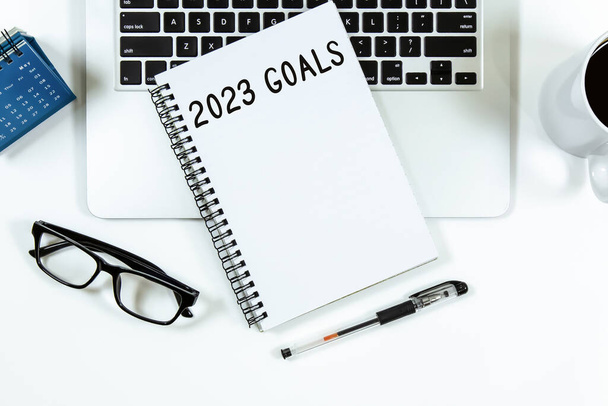 Top view of notebook handwriting goals for 2023 year, laptop and eyeglasses on white background. Business plan concept. - 写真・画像