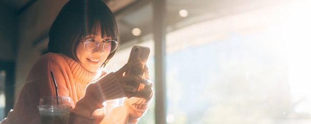 Young adult happy asian woman wear eyeglasses using mobile phone for social media application. Text online message at indoor cafe on day. Banner size background with copy space. - Фото, изображение
