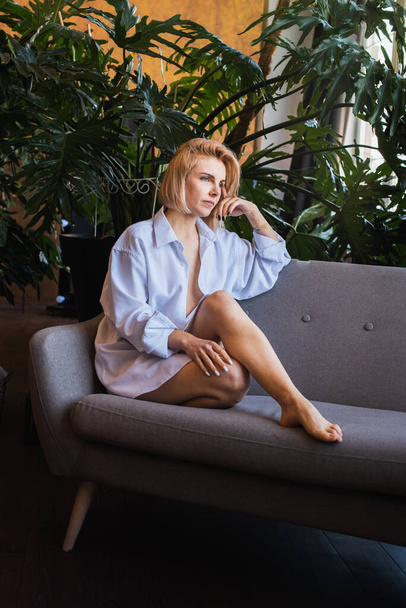 young blonde, beautiful girl sitting on the couch by the window in a white shirt with tropical flowers, organic skin care, luxury spa hotel, lifestyle photo. Selective focus, defocus - Photo, Image