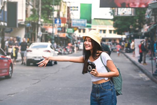 Joven viajero asiático mujer feliz en camisa blanca con mochila. Sostenga cámara instantánea. Marcar abajo de un taxi para el turismo. Fondo de la carretera Khaosan. Bangkok City, Tailandia - Foto, Imagen