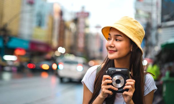 Young asian solo traveler woman in white shirt with backpack. hold instant camera beside landmark Yaowarat. Chinatown street food bokeh background with copy space. Bangkok City, Thailand - Photo, Image