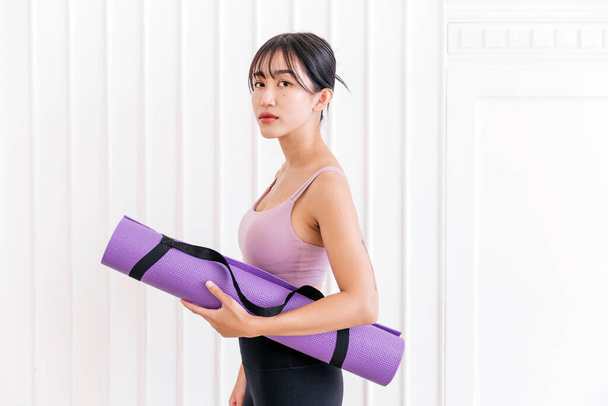 Asian woman wearing sportswear and yoga pants carrying a rolled-up yoga mat standing in front of white wall background with sunlight from the window. Half-length image with copy space. - 写真・画像