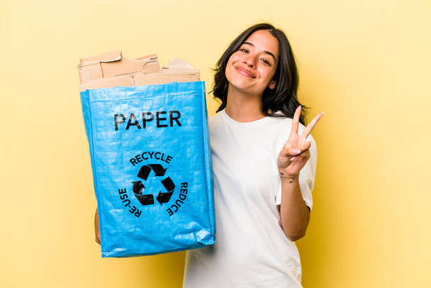 Jovem hispânica reciclando papel isolado em fundo amarelo alegre e despreocupado mostrando um símbolo de paz com os dedos. - Foto, Imagem
