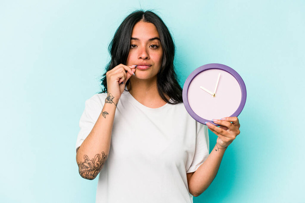 Young hispanic woman holding a clock isolated on blue background with fingers on lips keeping a secret. - Foto, imagen